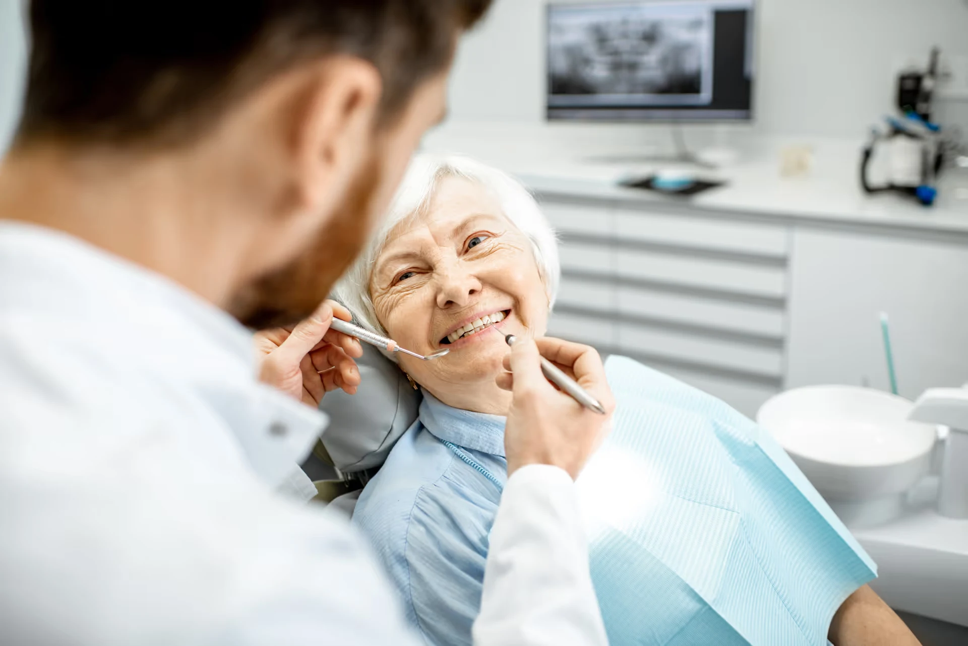 Dentist examining an older female patient, both smiling, x-ray visible on monitor